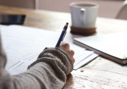 Woman's hand writing on paper with notebook and coffee mug in background. Photo by Unseen Studio on Unsplash